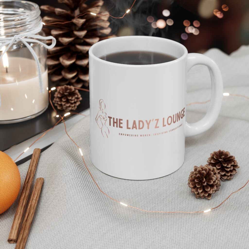 White ceramic mug with "THE LADY'Z LOUNGE" logo in rose gold, featuring a silhouette of a woman. The mug is filled with coffee, surrounded by pinecones, fairy lights, a candle, and an orange, creating a cozy and empowering scene.